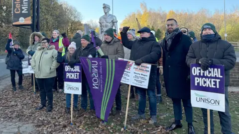 Zoe Head-Thomas/PA Wire About 15 people standing on a picket line, with Official Picket - Unison signs. They are wearing coats, hats and scarves and are standing in front of a statue of a miner wearing a helmet with his sleeves rolled up.