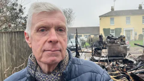 Andrew Turner/BBC Ron Anderson looking into the camera while wearing a patterned scarf and blue coat. He is stood in front of burnt remains, with a yellow house on the opposite side of the street.