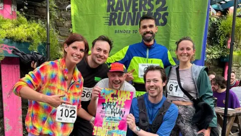 Six runners smiling at the camera in bright shirts and t shirts and tops. They are in front of a Ravers 2 Runners neon green banner and holding a leaflet for Dry Run. Behind them, there are some poeple sitting at picnic benches.