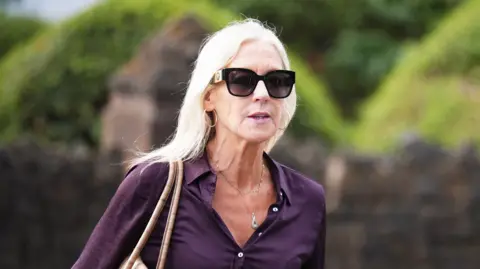 A woman with blonde hair is wearing a purple shirt and sunglasses and hoop earrings.