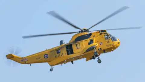 Royal Air Force / Ministry of Defence A yellow helicopter is flying through the blue sky. On the side there is writing which says Royal Air Force Rescue and the tail number XZ594 is visible. There is a person standing in the open side door wearing a yellow helmet.