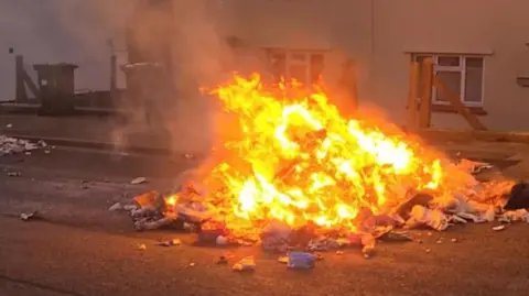 A mound of rubbish burns brightly in the middle of a road in front of houses and wheelie bins.