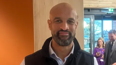 The man is standing in a bright, modern room with wooden beams and glass doors, wearing a light blue shirt and a dark vest. Several people are gathered and talking behind him, giving the scene the feel of a formal or commemorative event.