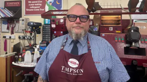 Kenny Bold is in a Timpson shop wearing his Timpson apron and a check shirt. Equipment used in his shop for shoes and keys is behind him.