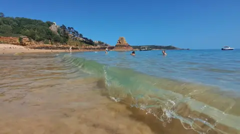 A few people are swimming in a blue sea at Les Creux. There are several small boats off the coast and the sky is blue and cloudless. In the distance several people can be seen relaxing on the sandy beach.