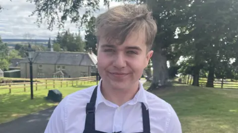 Person wearing a white shirt and a dark navy apron with a “Highland Farm Café” logo, standing on a paved path in a rural outdoor setting. Behind the person are green lawns, wooden fences, mature trees, and a stone building, with a cloudy sky and countryside visible in the distance.