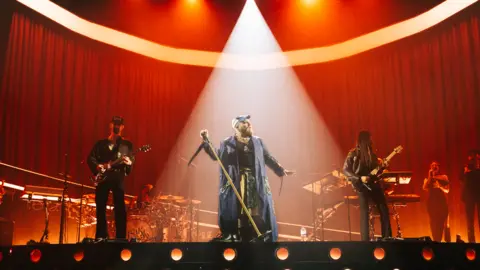 Kyle Horne Teddy Swims performing a concert. He is standing on stage with a spotlight on him. There are other band members who are cast in the red stage lights. Teddy is holding the microphone stand to his left side while looking to his right. He is wearing a baseball cap and long purple coat.