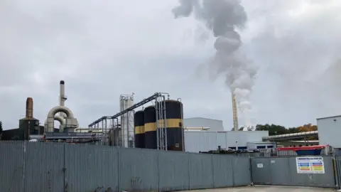 Charles Heslett/BBC Grey metal fencing with stainless steel chimneys and black and gold storage silos behind and while smoke coming from one building. 