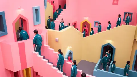 Netflix Contestants in dark blue track suits walk up and down the distinctive staircase which is painted pink, blue and yellow.
