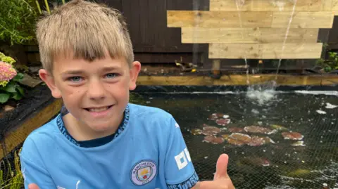 James smiles and gives two thumbs up to the camera. Behind him is a large pond, covered by netting, and several fish can be partially seen beneath the surface of the water.