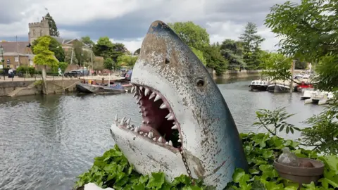 A large metal shark's head in the style of Jaws rearing up out of some foliage at the side of a river