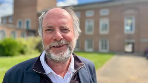 Liberal Democrat leader Julian Brazil standing in front of County Hall and wearing a white shirt, a maroon zip-through jumper and a navy blue blazer