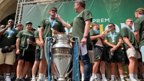 George, a man with short brown hair and a moustache, being interviewed on stage while other players look on from behind.
