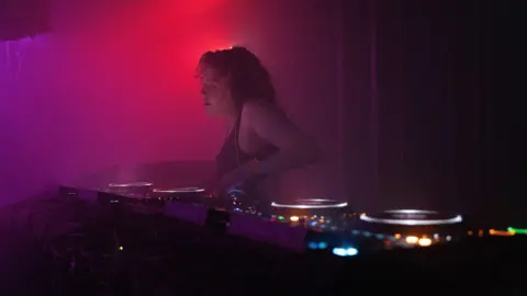 Jacinta Owen Jacinta stands behind DJ decks, she wears headphones