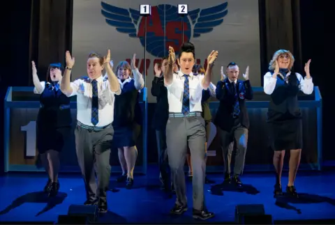PA Media Performers in airline-style uniforms dance in formation on stage during a scene from The High Life: The Musical, with a scoreboard-style set behind them.