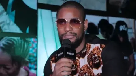 He is wearing a white tshirt with short black sleeves. The front has the repeated pattern of a roaring lion. He is wearing tinted sunglasses and is indoors. He is holding a mic.