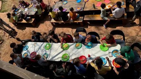 BBC Dozens of children sit at three long tables which are placed in the schoolyard. They are eating lunch from brightly coloured plastic plates. The picture is taken from above, so you can only see the tops of their heads