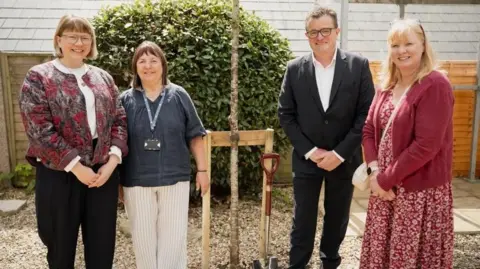 Three women and a man standing around a cherry tree and a shiny spade