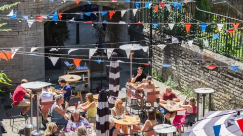 Diners eat in a sunny restaurant garden. Red, white and blue bunting is hanging above them.