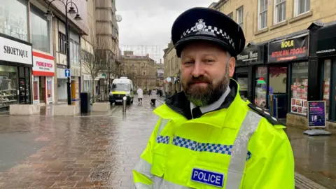 A bearded man wearing a police hat and fluorescent jacket standing on a street