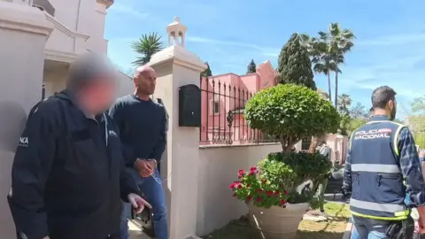National Crime Agency/PA Wire Steven Bullen walking out of a white-walled gate wearing a dark sweatshirt, blue jeans and with his cuffed hands in front of him. On the right is man with a blurred out face wearing a jacket labelled NCA (National Crime Agency) and on the left is the back view of a Spanish officer with policia nacional written on the back of his blue jacket. 