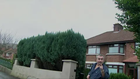 BBC Freddie Scappaticci pointing at the camera with his eyebrows raised. Semi-detached houses and a hedge are visible behind him.