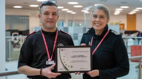 Scott Fitzsimmons standing next to Nikki Watson, they are both holding a certificate in a frame. They have red lanyards round their necks and are wearing black tops. Scott has short dark hair and glasses on his head and Nikki has tied back long hair. She is smiling. They are inside a building, which is slightly blurred behind them.