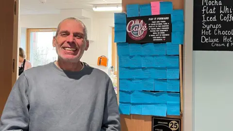 A man smiling straight at the camera standing next to a notice board. It is covered in blue sticky notes and has the words "Pay it forward. Times are tough. If you're in need, take a post-it to the counter. If you're able, consider paying for someone's meal or drink. A small act of kindness can make a big difference" at the top.