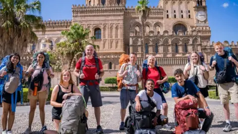 BBC/Studio Lambert A group of people with backpacks pose smiling in front of a big sandstone building. They are dressed for warm weather and there are palm trees in the background