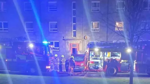 Fubar News Firefighters next to fire engines in darkness with their blue lights shining.