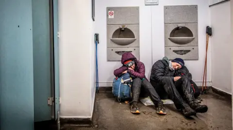 Two runners who are caked in mud and are wet sit slumped on the floor of public toilets. They are sleeping on their bags and look very dishevelled. 