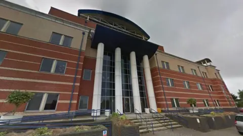 Google A red brick building entrance with four white columns by its glass frontage.