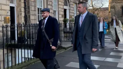 David Toshack, right, wearing a dark grey suit over a white shirt and a pink tie arriving at the employment tribunal.
