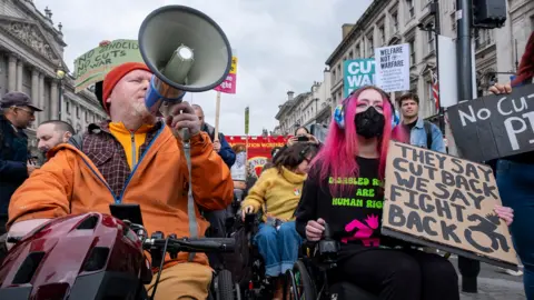 People in wheelchairs are pictured at a demonstration in central London with various signs, the most prominent of which reads 'they say cut back we say fight back' and 'welfare not warfare', and one of the protesters is holding up a megaphone. There are also other people standing behind them protesting as well and the sky is cloudy. Taken in London on 26 March.