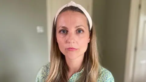 Julie Imrie looking straight ahead with a neutral expression. She has long, dark blonde hair and is wearing a white headband and a green shirt with blue on. 