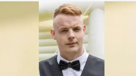 A head and shoulders shot of a young man wearing a black bowtie and waistcoat. He has ginger hair cut short.