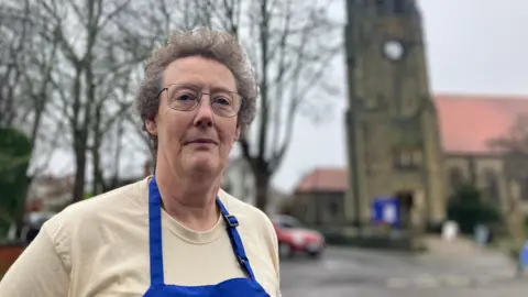 Elizabeth Baines/BBC A woman with short brown hair stands in front of a church. She wears a blue apron.