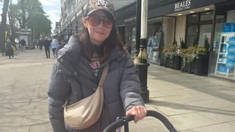 Megan Pennington, 33, who has a baseball cap with a NY logo, large framed dark red sunglasses, and a grey puffer coat, smiles at the camera while pushing the handle of a pram. 