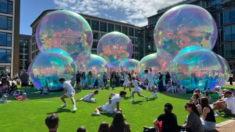 A large outdoor art installation that looks like giant bubbles, on a green floor that resembles grass. There is a dance performance happening in front of it. The dancers are wearing white t-shirts with white shorts or skirts. There is a crowd of people all around watching. 