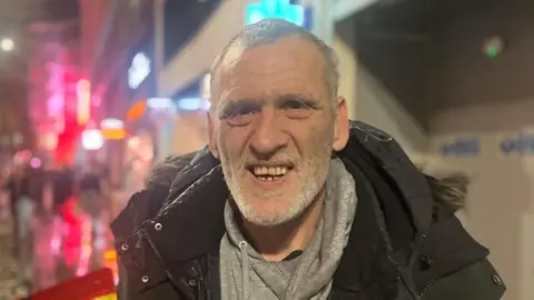 BBC Photograph of a homeless man called Gary O'Donnell from Liverpool. He is pictured by the Arndale Centre in Manchester city centre. He has grey short hair and a grey beard. He wears a grey hoodie and black parka jacket. He is smiling.