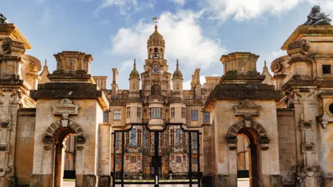 Harlaxton Manor The picture shows a gothic style building with a turret in the middle. It is made of stone and there are lots of towers either side with rectangular windows. There is a black iron gate at the bottom of the photo. 