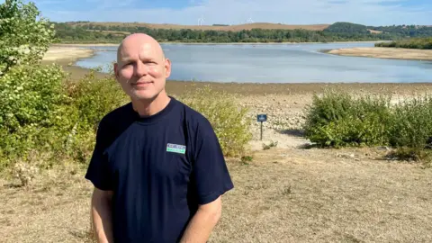 Doug Clarke, wearing a navy blue t-shirt with a Severn Trent logo, stands in front of Carsington Water.