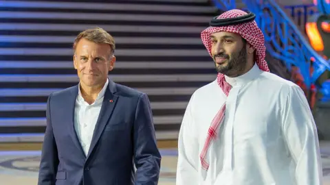 Esports Foundation Emmanuel Macron and Prince Mohammed Bin Salman (MBS) stroll side-by-side into the venue of the EWC closing ceremony. Macron wears a navy blue suit, over a white shirt, collar open and no tie. MBS wears a traditional thawb (long, white robe) and red and white head-dress.