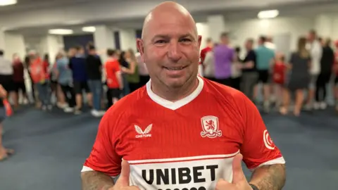 Russell Willis is wearing a red and white Middlesbrough top and has both thumbs up while smiling at the camera. He is bald and has tattoos up the length of his forearms. There is a line of people waiting in the airport behind hi, but they are out of focus.