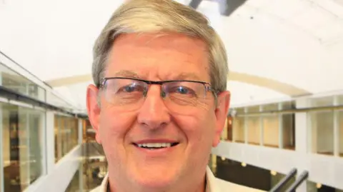 Peter Ashton A man smiles at the camera. He has short grey hair and wears glasses with a white shirt. 