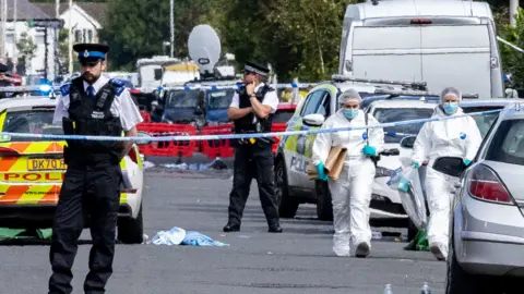 Police and forensic investigators in white protective clothing and masks are seen walking on a road filled with police vans and cars, with debris strewn across it and sealed off by blue and white police tape.