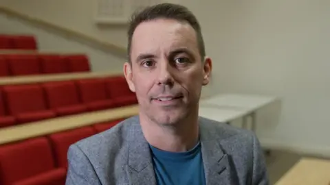 The principal of Dundee and Angus College Simon Hewitt. He has short black hair and brown eyes. He is wearing a grey suit jacket and light-blue tshirt 