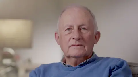 A man with short-cut grey hair and green eyes is looking directly at the camera. He can be seen, from the chest up, wearing a blue jumper and a shirt. Behind him, out of focus, is a small table and a lamp, in a dimly lit room with plain grey walls. 