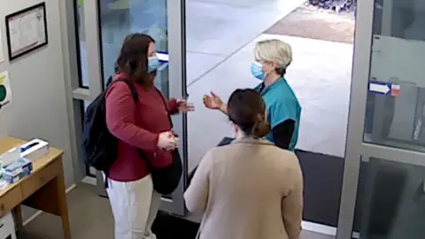 Three women stand talking in front of a sliding door at a hospital while wearing blue medical masks.