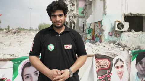 Red Crescent rescue worker Reza Besharati, pictured in a black T-shirt with Red Crescent badges on it. He is standing in front of banners showing the faces of people killed in the attack on the school, with the damaged buildings behind him.